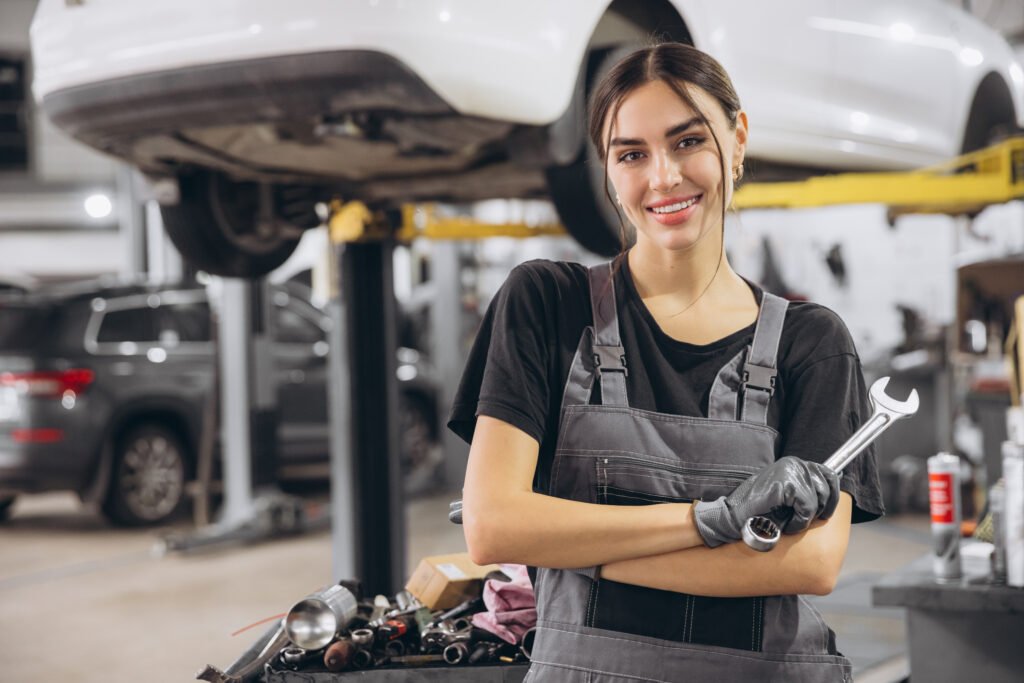 Smiling young woman mechanic standing beneath lifted car, holding tools and looking at camera in car service garage