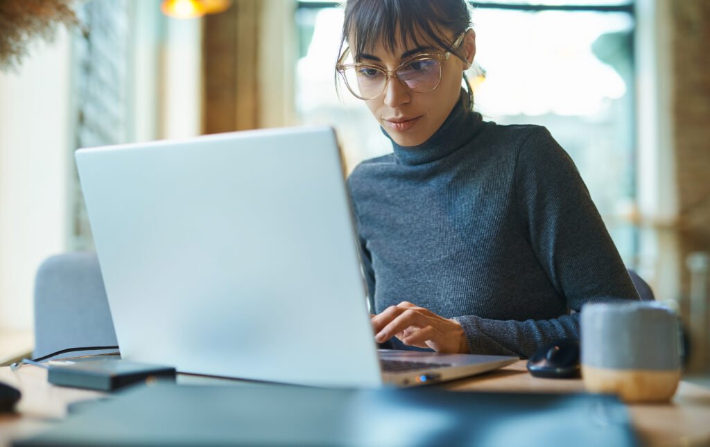 portrait of skilled young female developer in eyeglasses concentrating on screen and typing on laptop while sitting at desk at workplace or cafe.