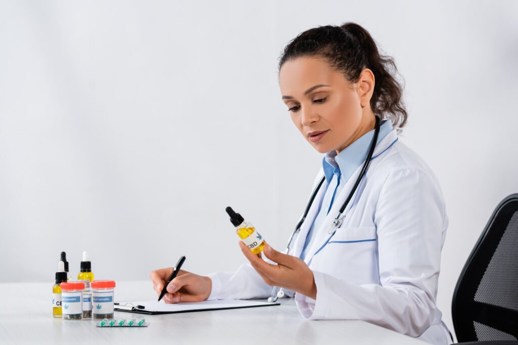 african american doctor writing prescription and holding cbd bottle near medication on desk