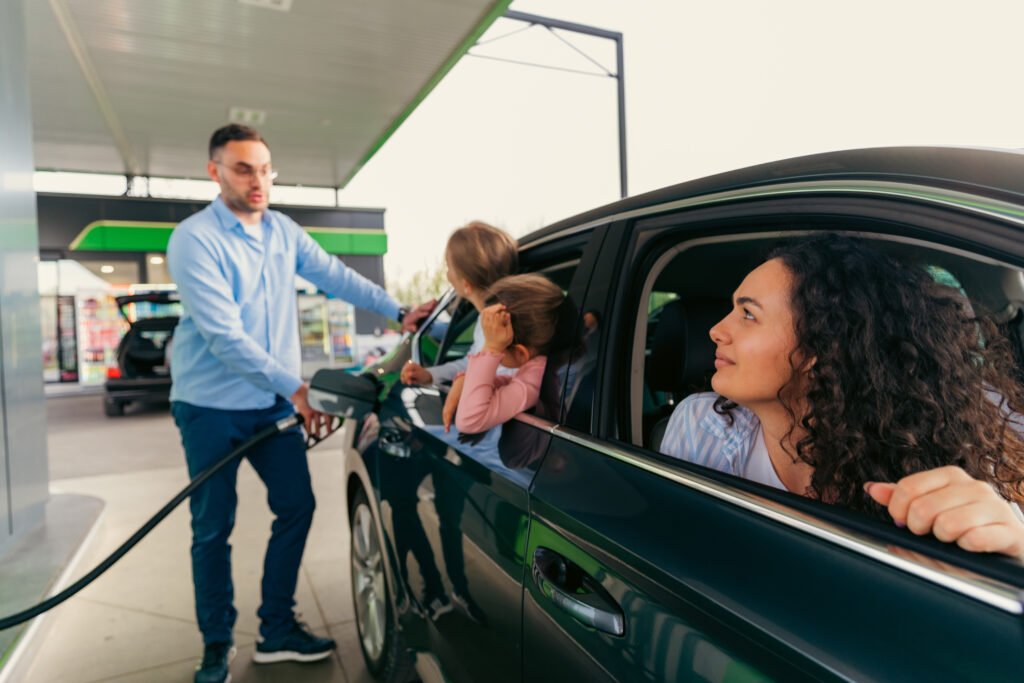 The father fills the fuel and talks to his daughters who are sitting in the back seat of the car, he is upset because they did unfasten their seat belts. Mom and daughters look at father